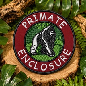 May include: A circular embroidered patch featuring a dark red border with the white text PRIMATE ENCLOSURE. The center displays a green background with a black and white illustration of a gorilla walking on all fours in a jungle setting.