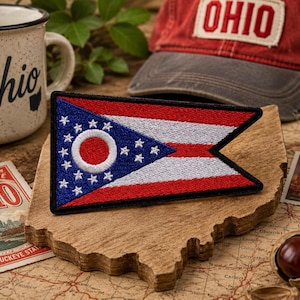 Può includere: Una toppa ricamata triangolare con il design della bandiera dello stato dell'Ohio, caratterizzata da un triangolo blu, stelle bianche, un cerchio bianco e strisce orizzontali rosse e bianche, bordata in filo nero su una sagoma in legno.