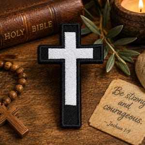 May include: A black and white embroidered cross patch featuring a central white cross shape set against a black rectangular background with a stitched border, displayed alongside a wooden rosary, a Bible, a candle, and a stone with text.