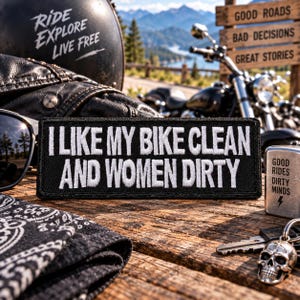 May include: A rectangular black embroidered patch featuring white text that reads I LIKE MY BIKE CLEAN AND WOMEN DIRTY. The patch has a stitched border and is positioned on a wooden surface alongside a metal lighter and a skull keychain.