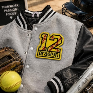 Könnte beinhalten: Graue Collegejacke mit schwarzen Lederärmeln und einem gelben Aufnäher mit der Nummer 12 und dem Namen Braelyn. Ein Softball, ein Handschuh und ein Helm sind ebenfalls zu sehen.