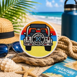 May include: A round patch featuring a jeep with the text "Wander Often" in the center. The patch has a colorful sunset design in the background. The patch is on a piece of driftwood, with a straw hat, sunglasses, and a water bottle in the background.