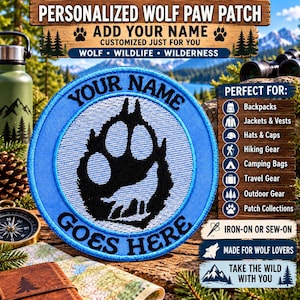 May include: A circular embroidered patch featuring a black wolf paw print on a light blue background. The text YOUR NAME GOES HERE is stitched in black around the paw print. The patch has a blue border and is designed for iron-on or sew-on application.