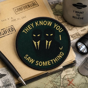 May include: A circular embroidered patch with a dark green background and a black border. It features two silhouetted figures in suits with yellow eyes and yellow text that reads "THEY KNOW YOU SAW SOMETHING" in a circular arrangement.