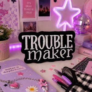 Puede incluir: Un parche negro con texto bordado blanco que dice "TROUBLE maker". La imagen también presenta un cuaderno morado, una planta en maceta rosa, una bola de discoteca y una estrella de neón. Otros elementos incluyen bolígrafos, libros y pegatinas decorativas.
