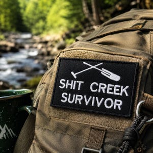 May include: A black patch with white text and crossed oars that reads "SHIT CREEK SURVIVOR" is attached to a brown backpack. A green and white speckled camping mug is visible in the background, with a river and trees.