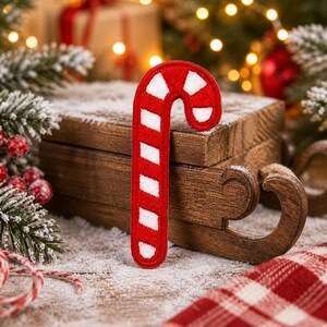 以下が含まれることがあります： 赤と白のキャンディケーンパッチ、お祝いのクリスマスの装飾が、人工雪をまぶした木製の表面に置かれています。 キャンディケーンは、赤と白のストライプが特徴の、古典的な休日のシンボルです。 木製のそりや赤と白のチェック柄の布も見えます。