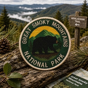 May include: A circular embroidered patch featuring a black bear silhouette against a green mountain landscape and a yellow sky. The text GREAT SMOKY MOUNTAINS NATIONAL PARK is stitched in white around the border with a tan edge finish.