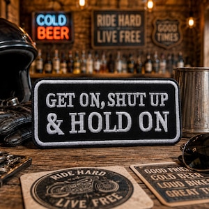 Könnte beinhalten: Ein schwarz-weißer Aufnäher mit dem Text "GET ON, SHUT UP & HOLD ON" liegt auf einer Holzoberfläche. Weitere Gegenstände sind ein Motorradhelm, Handschuhe, ein Metallbecher und eine Sonnenbrille. Schilder zeigen "COLD BEER", "RIDE HARD LIVE FREE" und "GOOD TIMES".