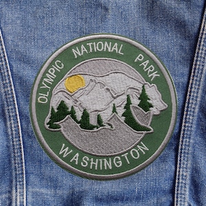 May include: Embroidered patch featuring the Olympic National Park in Washington. The circular patch has a green border with white text and a central design of mountains, trees, and a bear. A yellow sun is also visible.