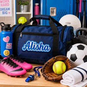 K&ouml;nnte beinhalten: Eine marineblaue Sporttasche mit dem Namen Alisha in wei&szlig;er und blauer Schreibschrift auf der Vorderseite. Die Tasche hat schwarze Griffe und Rei&szlig;verschl&uuml;sse und ist umgeben von einer blauen Trinkflasche, pinken Fu&szlig;ballschuhen, einer Trillerpfeife, einem Baseballhandschuh, einem Ball und einem Handtuch.