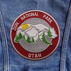 May include: Embroidered patch featuring the Zion National Park in Utah. The circular patch has a red border with the words "ZION NATIONAL PARK UTAH". The design includes a mountain range, trees, and a yellow sun.