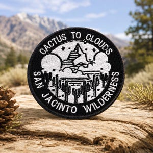 Puede incluir: Un parche bordado circular con fondo negro y costuras blancas. El diseño muestra un pico de montaña, nubes y cactus saguaro con el texto CACTUS TO CLOUDS SAN JACINTO WILDERNESS dispuesto en un círculo alrededor del borde del parche.