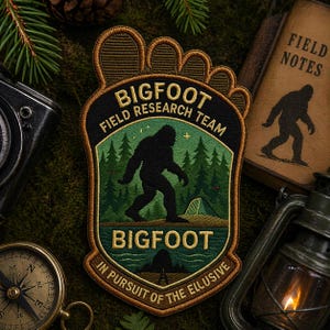 May include: Embroidered Bigfoot Field Research Team patch with a black silhouette of Bigfoot walking in front of a forest and a tent. The patch has a foot-shaped border and text that reads "Bigfoot Field Research Team" and "In Pursuit of the Elusive."