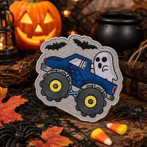 May include: A gray embroidered patch featuring a blue monster truck with a spiderweb design on the side. A white ghost sits in the truck bed, and two black bats are stitched above the vehicle. The patch has a scalloped edge and gray background.