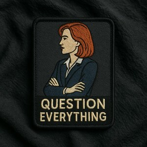 May include: A rectangular embroidered patch with a black background featuring a person with shoulder-length auburn hair wearing a dark blue blazer over a white collared shirt with arms crossed. The text "QUESTION EVERYTHING" is in beige below.