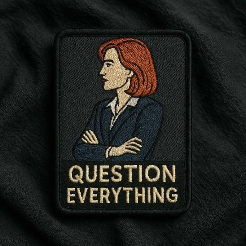 May include: A rectangular embroidered patch with a black background featuring a person with shoulder-length auburn hair wearing a dark blue blazer over a white collared shirt with arms crossed. The text "QUESTION EVERYTHING" is in beige below.