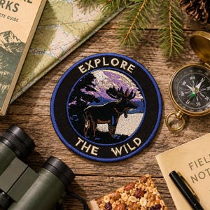 May include: Circular embroidered patch featuring a moose design with the text "EXPLORE THE WILD". The patch is black, blue, and white, and is on a wooden surface with a compass, binoculars, and a map.