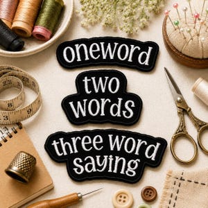 Op de afbeelding: Drie zwarte geborduurde patches met witte tekst die oneword, two words en three word saying leest. De patches zijn omringd door naaibenodigdheden, waaronder een meetlint, garenklosjes, een schaar, een vingerhoed en spelden.