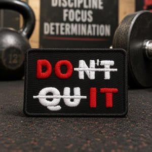 May include: Black rectangular patch with the words "DON'T QUIT" embroidered in red and white. The words are crossed out with a white line. The patch is on a textured surface, with a kettlebell and weight plate in the background.