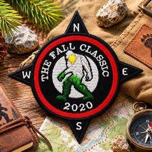 May include: A circular embroidered patch shaped like a compass rose with black, red, and white thread. The center features a white Bigfoot figure with a yellow sun, green legs, and the text THE FALL CLASSIC 2020. Cardinal directions N, S, E, W are stitched in white on the black points.