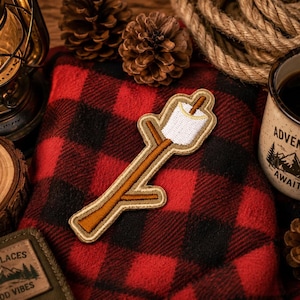 May include: A patch featuring a marshmallow on a stick, with a brown and tan color scheme, rests on a red and black plaid blanket. A mug with the words "Adventure Awaits" and a camping scene is nearby, along with a lantern and pine cones.