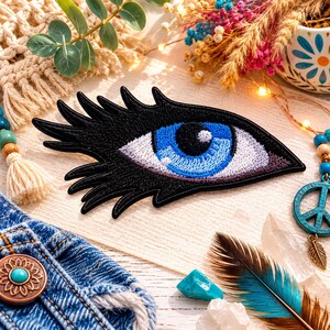 May include: An embroidered patch shaped like an eye with long black eyelashes, a bright blue iris, and a black pupil. The design features detailed stitching in shades of black, white, and blue, creating a distinct graphic representation of an eye.