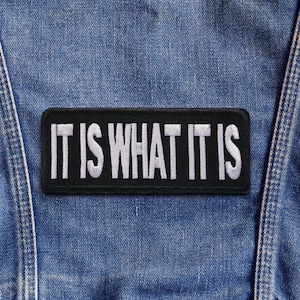 May include: A black rectangular patch embroidered with the words "IT IS WHAT IT IS" in white. The patch is attached to a blue denim jacket, with the jacket's seams and stitching clearly visible.