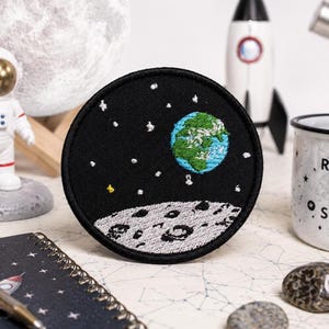 Puede incluir: Un parche bordado circular con fondo negro, estrellas blancas, una pequeña estrella amarilla, una Tierra azul y verde, y una superficie lunar gris con cráteres en la parte inferior. El parche tiene un borde cosido de color negro.
