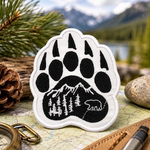 Pode incluir: Um emblema bordado em branco e preto com o formato de uma pegada de urso. A almofada central apresenta um gráfico branco detalhado de uma cordilheira, uma floresta de pinheiros e a silhueta de um pequeno urso sobre um fundo preto.