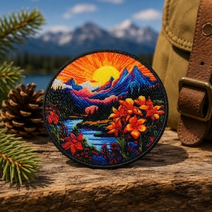 Könnte beinhalten: Runder, gestickter Aufnäher mit einer Berglandschaft mit Sonnenuntergang, blauen Bergen und orangefarbenen Blumen. Der Aufnäher hat einen schwarzen Rand und liegt auf einer Holzoberfläche.