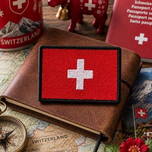 Op de afbeelding: Een rechthoekige geborduurde patch met de Zwitserse vlag, bestaande uit een wit kruis in het midden op een effen rode achtergrond. De patch heeft een dikke zwarte rand en ligt op een notitieboek met een bruine leren kaft.