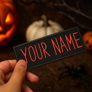 Puede incluir: Un parche rectangular negro con las palabras "YOUR NAME" bordadas en rojo. El parche se sostiene en una mano, con un fondo borroso con temática de Halloween con calabazas y una araña.