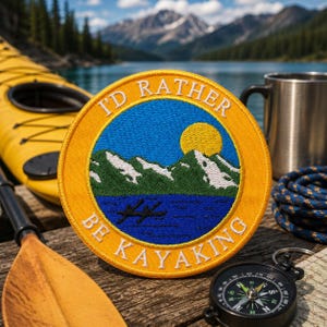 Könnte beinhalten: Ein runder gestickter Aufnäher mit gelbem Rand und dem weißen Text I'D RATHER BE KAYAKING. Die Mitte zeigt einen blauen See, grüne Berge mit weißen Schneekappen, eine gelbe Sonne und zwei schwarze Silhouetten von Kajaks auf dem Wasser.