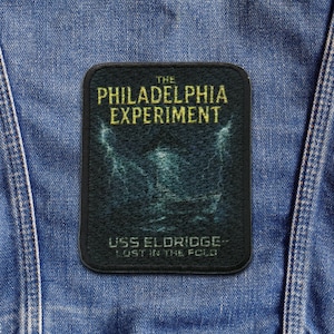 Op de afbeelding: Een zwart rechthoekig embleem met de tekst "THE PHILADELPHIA EXPERIMENT" in gouden letters. Daaronder een vage afbeelding en de tekst "USS ELDRIDGE - LOST IN THE FOLD". Afgeronde hoeken.