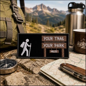 May include: A rectangular black embroidered patch featuring a white stick figure hiker and a brown sign with the text YOUR TRAIL YOUR PARK and MILES. The patch is positioned on a textured surface with a compass, a map, and a metal water bottle.