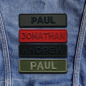 May include: Four rectangular embroidered patches with names on a denim jacket. The patches are in dark green, red, black, and olive green. The names are "PAUL", "JONATHAN", "ANDREW", and "PAUL".