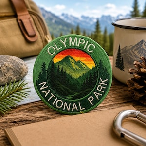 Puede incluir: Un parche bordado circular que presenta un paisaje de una cadena montañosa verde bajo un cielo de atardecer naranja y amarillo vibrante, rodeado por un borde verde oscuro con el texto blanco OLYMPIC NATIONAL PARK cosido a lo largo del borde.