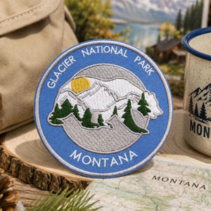 May include: A circular embroidered patch featuring a blue border with the white text Glacier National Park and Montana. The center depicts white snow-capped mountains, a yellow sun, and dark green pine trees against a light gray background.