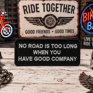 May include: Black rectangular patch with white embroidered text: "NO ROAD IS TOO LONG WHEN YOU HAVE GOOD COMPANY." The patch is surrounded by biker-themed decor, including a neon sign, a skull, and a motorcycle sign.