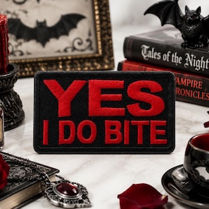 May include: Black patch with red embroidered text reading "YES I DO BITE." The patch is rectangular and has a black border. The image also includes a red candle, a small bottle labeled "Blood," and books with a bat figurine.