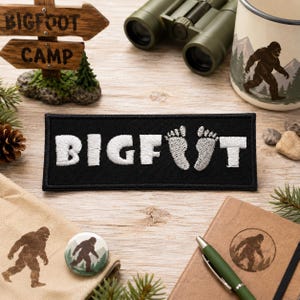 Könnte beinhalten: Ein rechteckiger, schwarz bestickter Aufnäher mit dem weißen Schriftzug BIGFOOT, bei dem die beiden Buchstaben O durch zwei weiße, gestickte Fußabdrücke ersetzt sind. Der Aufnäher hat einen schwarzen Stickrand und liegt auf einer Holzoberfläche.