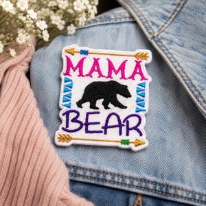 May include: Embroidered patch with the words "MAMA BEAR" in pink and purple, featuring a black bear silhouette. The patch is bordered with blue and gold arrow designs, set against a white background. The patch is on a denim jacket.