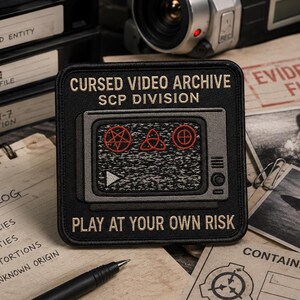 May include: A square black embroidered patch featuring a grey television set with static on the screen. The text reads CURSED VIDEO ARCHIVE SCP DIVISION and PLAY AT YOUR OWN RISK. Three red symbols are displayed on the screen above a white play icon.