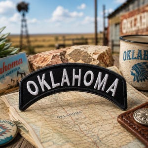 May include: A black embroidered patch with the word "OKLAHOMA" in white arched across the top. The patch is on a map of Oklahoma, with a vintage postcard and a mug with a Native American design. A leather wallet and a pin are also visible.