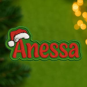 Könnte beinhalten: Gestickter Aufnäher mit dem Namen "Anessa" in Rot, umrandet in Grün, mit einem Weihnachtsmützen-Design. Der Aufnäher hat einen grünen Rand und einen weißen Weihnachtsmützenbesatz. Der Hintergrund ist ein verschwommenes Grün mit Bokeh-Lichtern.