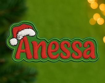 Könnte beinhalten: Gestickter Aufnäher mit dem Namen "Anessa" in Rot, umrandet in Grün, mit einem Weihnachtsmützen-Design. Der Aufnäher hat einen grünen Rand und einen weißen Weihnachtsmützenbesatz. Der Hintergrund ist ein verschwommenes Grün mit Bokeh-Lichtern.