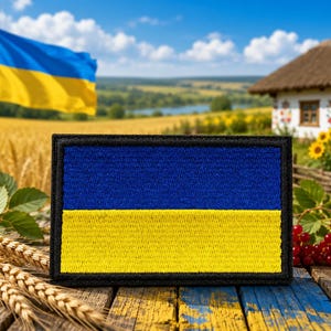 May include: A rectangular patch featuring the Ukrainian flag, with a blue upper half and a yellow lower half, framed by a black border. The patch is set against a backdrop of a wheat field, a traditional Ukrainian house, and a waving Ukrainian flag.