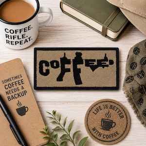 Peut inclure: Un écusson rectangulaire avec un fond en tissu beige et une bordure noire. Le mot COFFEE est brodé en noir, avec la lettre F en forme de fusil. L'écusson est placé parmi une tasse à café, un carnet et un dessous de verre en liège.