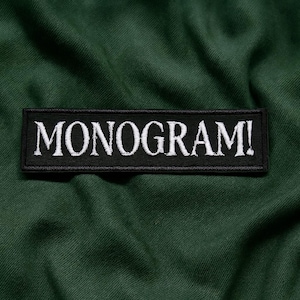 May include: A black rectangular patch with the word "MONOGRAM!" embroidered in white, set against a dark green fabric background. The patch is centered and the text is in a bold, stylized font.